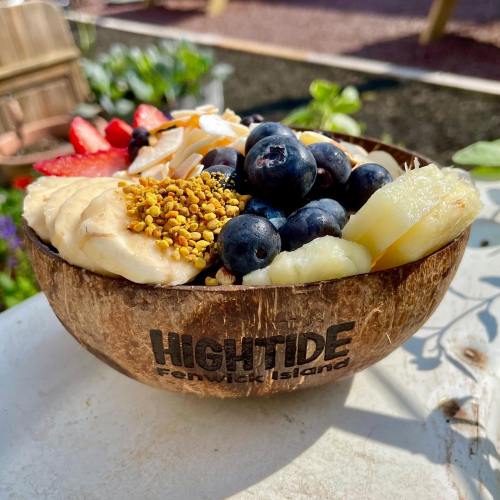 A coconut bowl filled with sliced bananas, blueberries, strawberries, and granola, set in a garden.