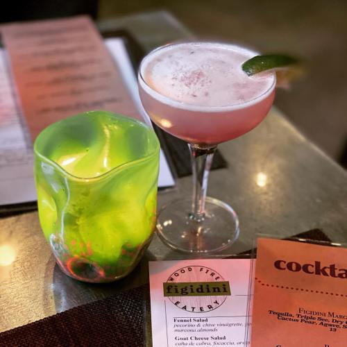 A vibrant cocktail with a lime garnish next to a colorful glass and a menu featuring a fennel salad.