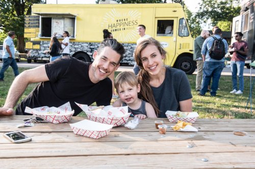 A smiling family of three sits at a picnic table with food, enjoying a sunny day at a food truck event.