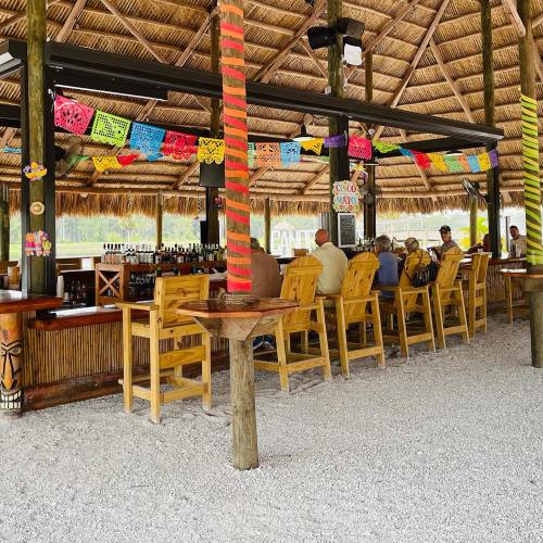 A tropical bar with wooden stools, colorful decorations, and a thatched roof, set in a sandy outdoor area.