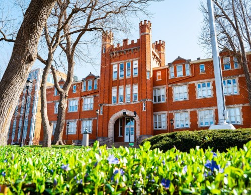 Historic red brick building with castle-like features, surrounded by green shrubs and blue flowers under a clear sky.