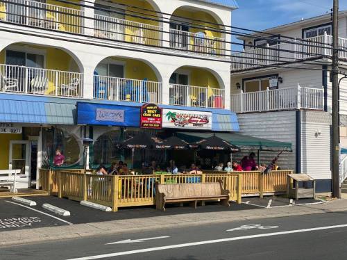 Outdoor dining area of a restaurant with yellow and white building, people seated under umbrellas, sunny day.