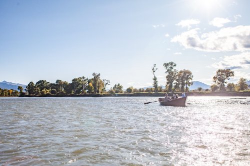 A small boat with people rows across a shimmering river, surrounded by trees and mountains under a clear blue sky.