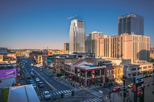 A vibrant cityscape featuring bustling streets, tall buildings, and lively storefronts under a clear blue sky.