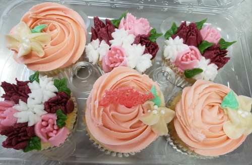 A clear plastic container holding six beautifully decorated cupcakes with floral designs in pink, white, and cream colors.