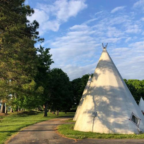 A scenic view of white teepees surrounded by green trees and a blue sky with scattered clouds.