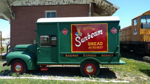 Vintage green delivery truck with a bright red sign advertising Sunbeam Bread, parked near a railway station.