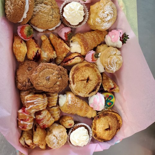 A colorful assortment of baked goods, including cookies, pastries, and cupcakes, arranged in a pink-lined box.