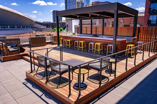 Rooftop terrace with wooden seating, a bar area, and city skyline in the background under a clear blue sky.