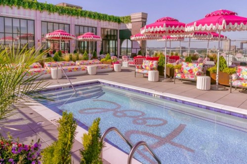 Rooftop pool area with colorful lounge chairs, umbrellas, and vibrant floral decor against a clear blue sky.