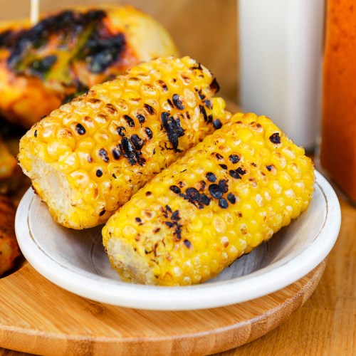 Grilled corn on the cob served on a small white plate, with charred kernels and a rustic wooden background.