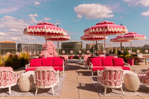 A vibrant rooftop lounge with pink umbrellas, plush seating, and city skyline views under a blue sky with fluffy clouds.