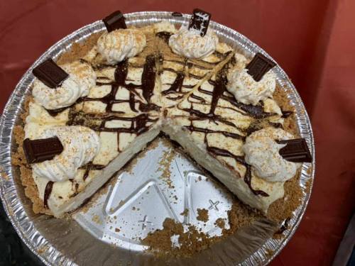A sliced cheesecake topped with whipped cream, chocolate drizzle, and chocolate pieces, in a silver pie dish.