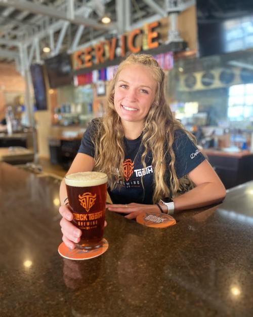 A smiling young woman holds a pint of beer at a bar, with a logo visible on her shirt and a lively background.