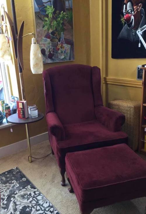 A cozy burgundy armchair with an ottoman, next to a lamp and a small table, in a warmly decorated room.