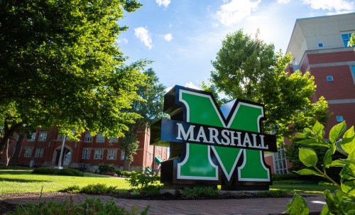 A large green and black "MARSHALL" sign in front of a university building, surrounded by trees and blue sky.