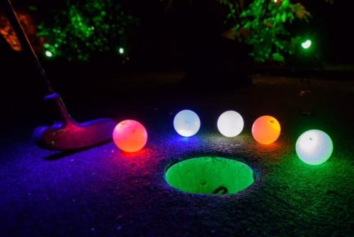 A putter beside glowing golf balls in various colors, with a hole illuminated in the background.