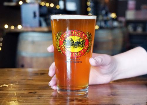 A hand holding a pint glass of amber beer with a "Firehouse Brewing Co." logo, set on a wooden table.
