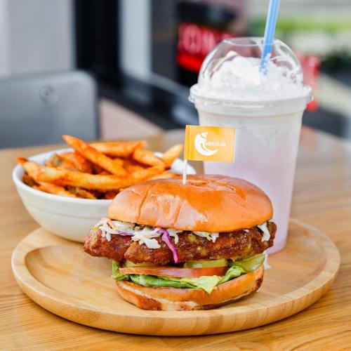 A crispy chicken burger with lettuce and slaw, served with sweet potato fries and a milkshake on a wooden plate.