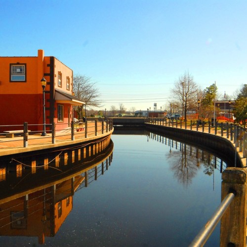 A calm canal reflects a colorful building under a clear blue sky, with trees lining the walkway.