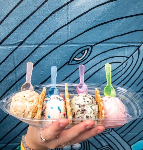 A hand holds a clear container with four colorful ice cream scoops and spoons against a blue mural background.