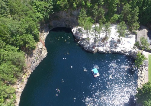 Aerial view of a serene swimming area with people in the water, surrounded by rocky cliffs and lush greenery.