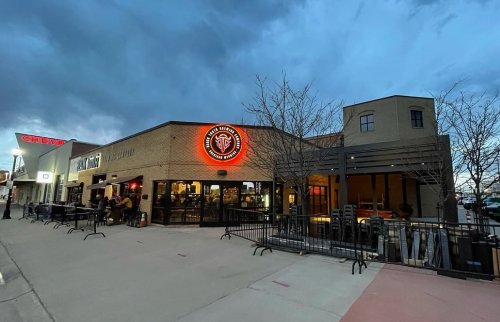 Exterior of a restaurant with a glowing logo, surrounded by trees and a cloudy sky, featuring outdoor seating.