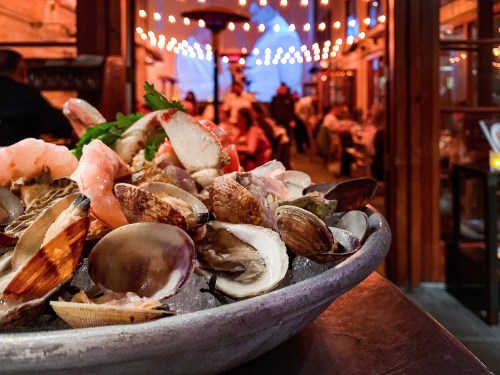 A platter of fresh seafood on ice, with a lively restaurant atmosphere and string lights in the background.