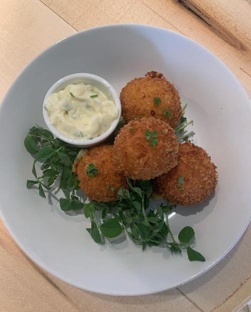 Four golden-brown croquettes on a plate with a small bowl of creamy dip and fresh herbs.