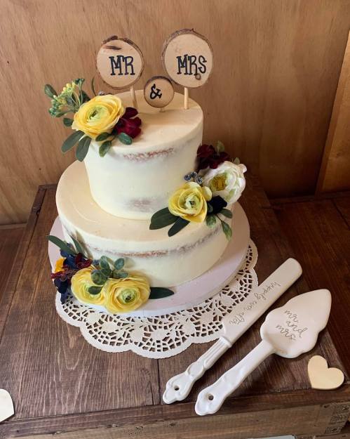 A two-tier wedding cake decorated with flowers and wooden "Mr & Mrs" signs, accompanied by a cake server set.