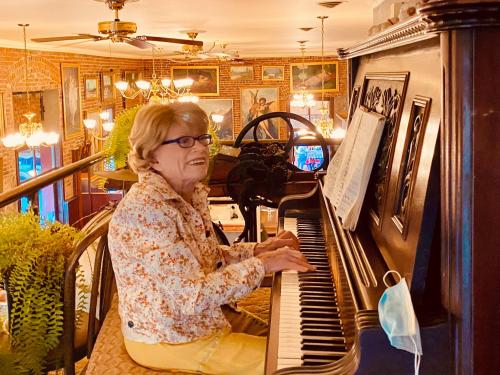 An elderly woman plays a piano in a cozy, well-decorated space with warm lighting and plants around her.