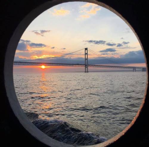 A sunset view of a bridge framed by a circular opening, with calm waters in the foreground and clouds in the sky.