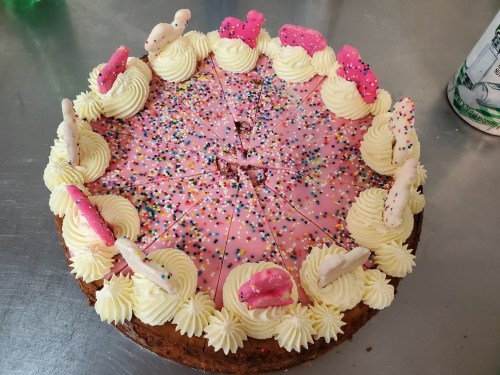 A round cake with pink frosting, whipped cream swirls, colorful sprinkles, and decorative cookies on top.