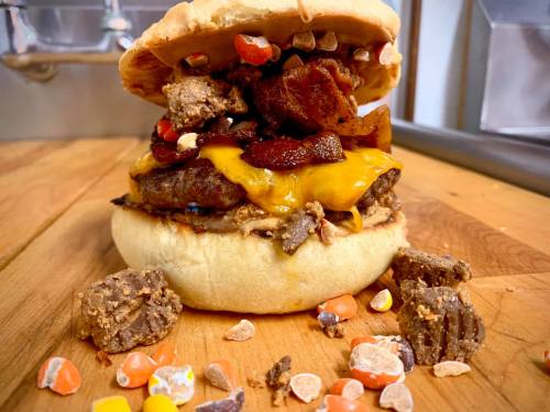 A towering burger with cheese, candy pieces, and toppings, resting on a wooden surface with scattered candy around it.