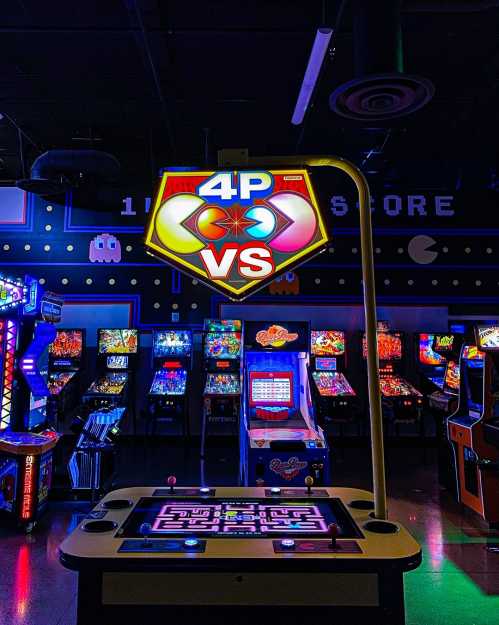 A brightly lit arcade sign reading "4P VS" above a game console, with vintage arcade machines in the background.