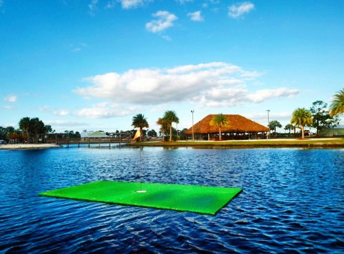 A floating green in a lake with a pavilion and palm trees in the background under a blue sky.