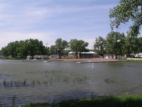 A serene lake scene with trees, a swimming area, and a building in the background under a clear blue sky.