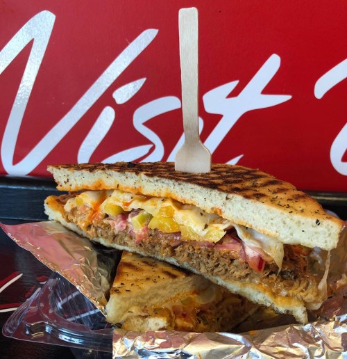 A grilled sandwich filled with meat, cheese, and vegetables, served on a foil wrapper against a red background.