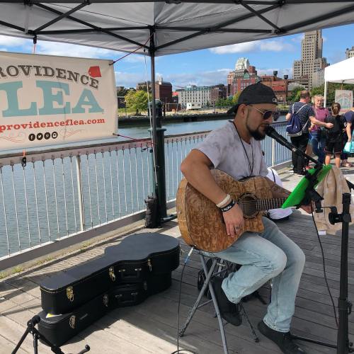 A musician plays guitar on a waterfront stage at an outdoor event, with a city skyline in the background.