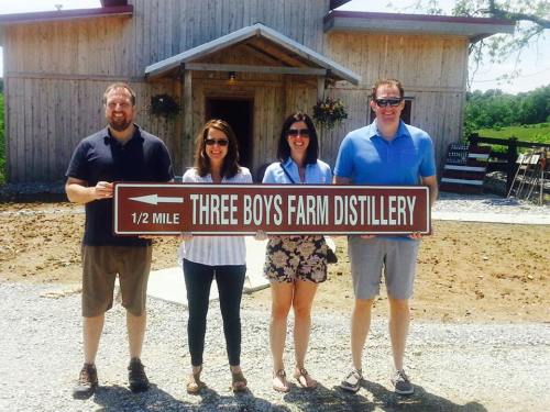 Four people stand in front of a wooden building, holding a sign that reads "Three Boys Farm Distillery 1/2 Mile."
