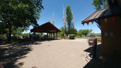 A sunny park scene featuring a pavilion, trees, and a playground in the background, with people enjoying the outdoors.