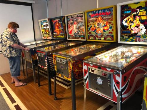A person in a floral shirt stands beside several colorful pinball machines in a retro arcade setting.