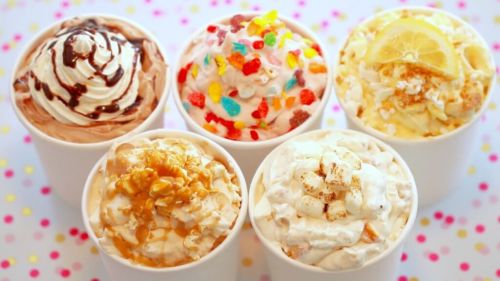 Five cups of colorful ice cream topped with whipped cream, sprinkles, and various toppings on a polka dot background.