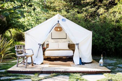 A cozy glamping tent with a bed, wooden chair, and lantern, set in a lush green outdoor environment.