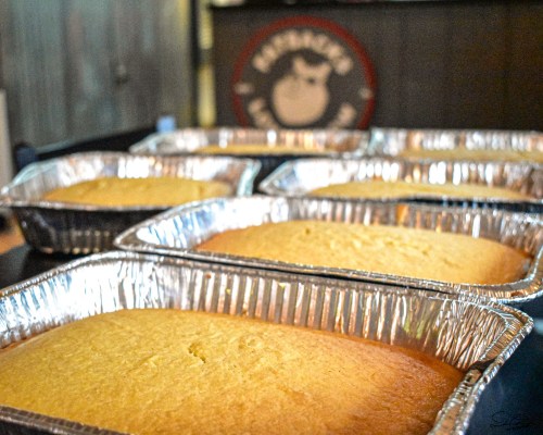 Six aluminum trays filled with freshly baked cakes, with a blurred background featuring a logo.