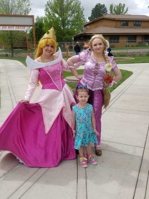 Two women in princess costumes pose with a young girl in a blue dress, smiling outdoors in a park setting.