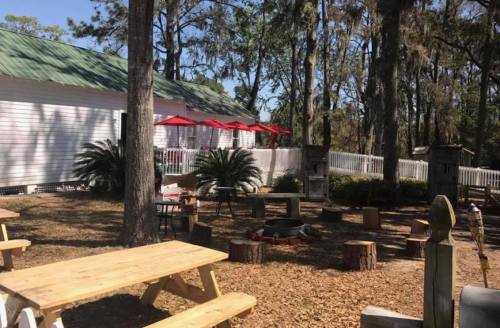 Outdoor seating area with picnic tables, fire pit, and shaded trees near a white building with red umbrellas.