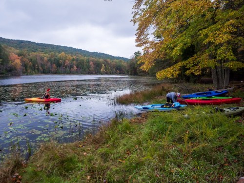 Two kayakers on a calm lake surrounded by autumn foliage, with colorful kayaks lined up on the shore.