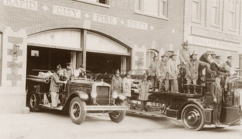 Historic fire department scene with firefighters posing on vintage fire trucks outside a brick fire station.
