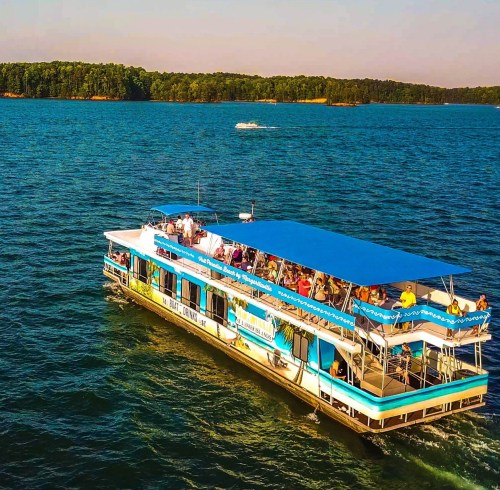 A colorful party boat with passengers cruising on a serene lake surrounded by green trees.